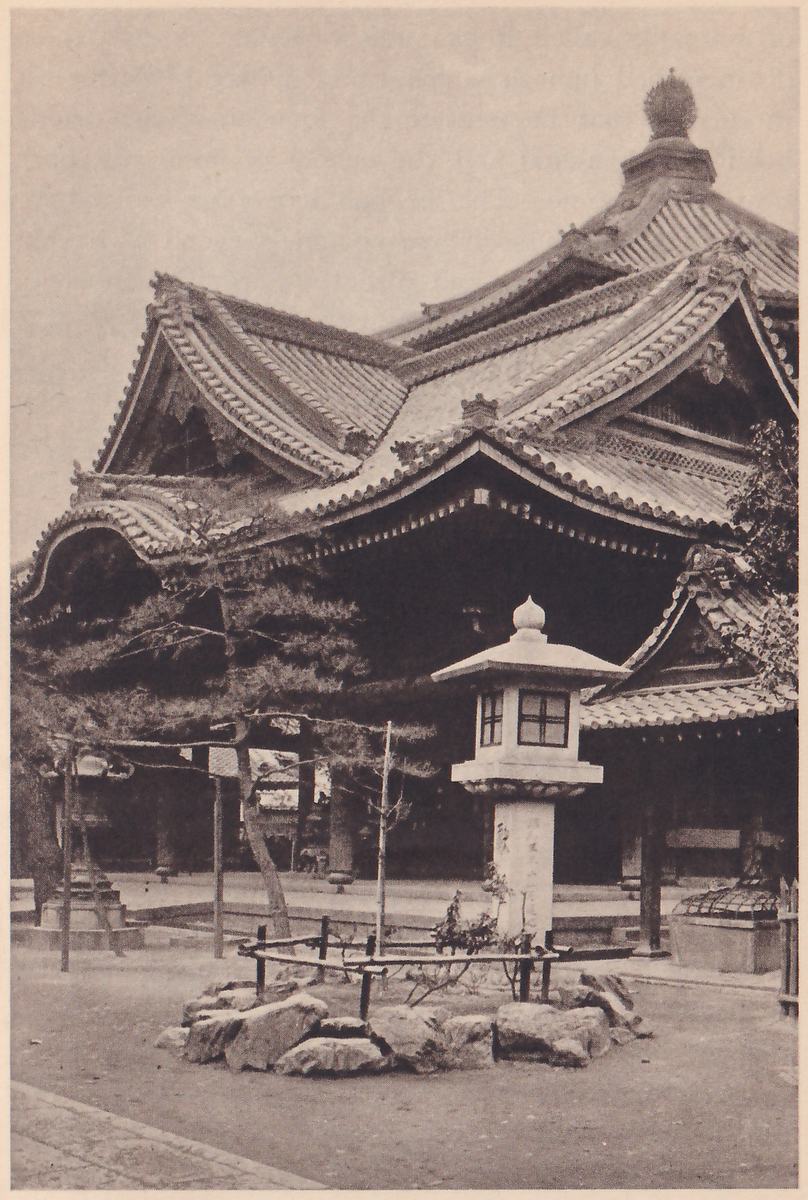 Photograph of a temple building with a tiered tiled roof topped by a finial, a pine tree and a stone lantern in front of it, and a ring of rocks on the ground in the foreground.