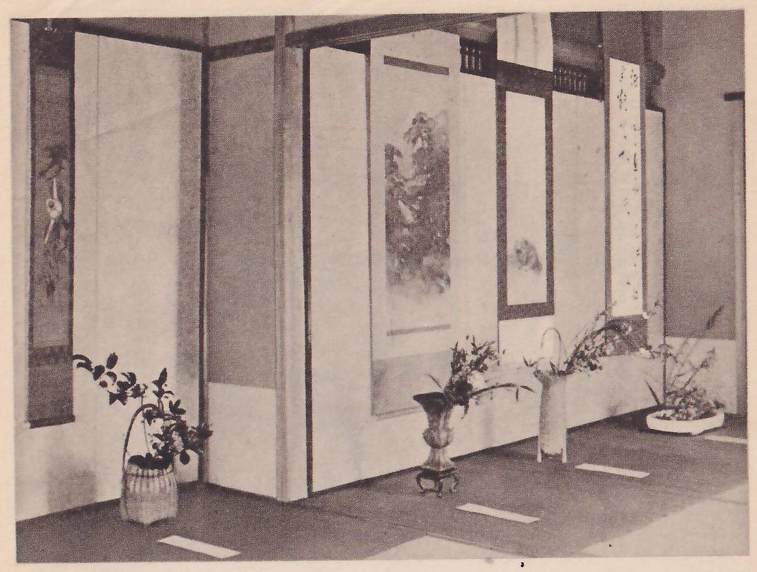 Photograph of a room with a long wall of panels on which four hanging scrolls hang, and on the floor in front of them four arrangements, in a basket, a footed bronze vase, a tall pale vase and a low white dish.