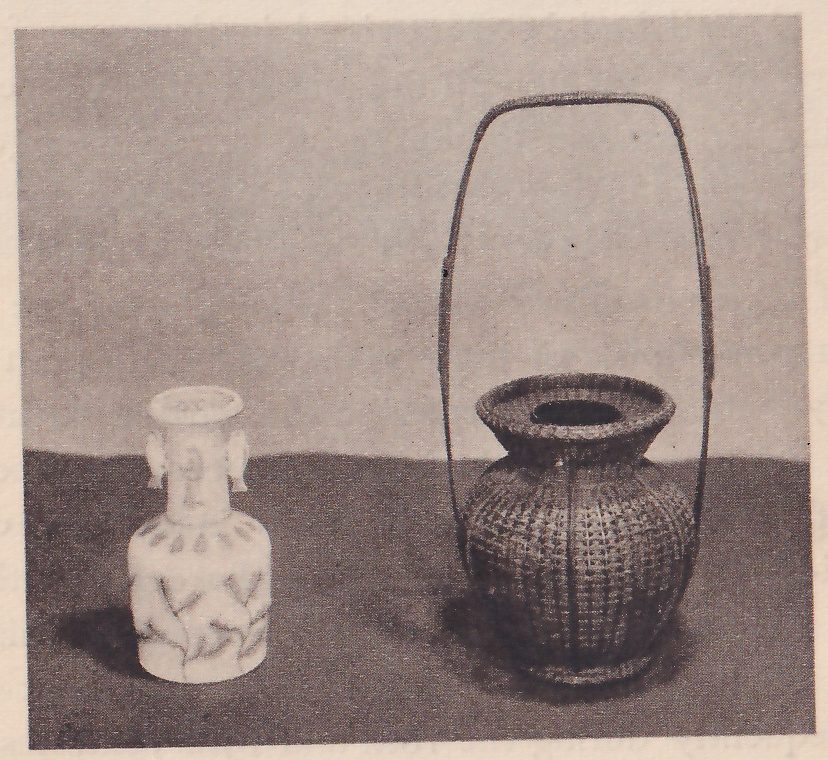Photograph of two vases on a dark ground: at the left a small pale china vase with a narrow neck, two small handles and a painted leaf pattern, and at the right a taller dark wicker basket with a wide mouth and a thin arched handle.