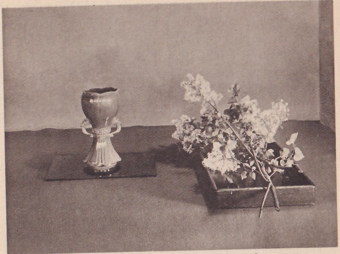 Photograph of a small footed vase with two handles on a flat dark mat at the left, and at the right a shallow dark tray with a spray of white flowers and leaves laid in it.
