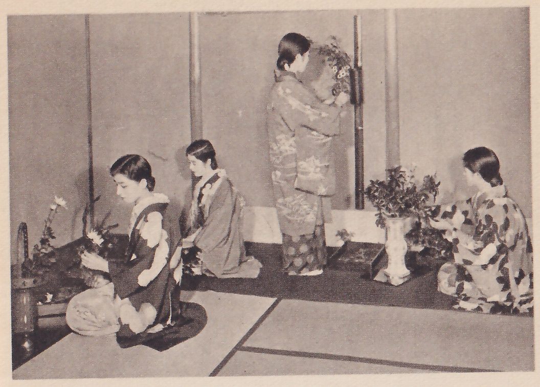 Photograph of four women in kimono in a mat room at an alcove: one kneels at the left working on flowers, a second kneels beside her, a third stands in the middle at the wall and reaches up to a hanging vase, and a fourth kneels at the right with a vase of leaves.