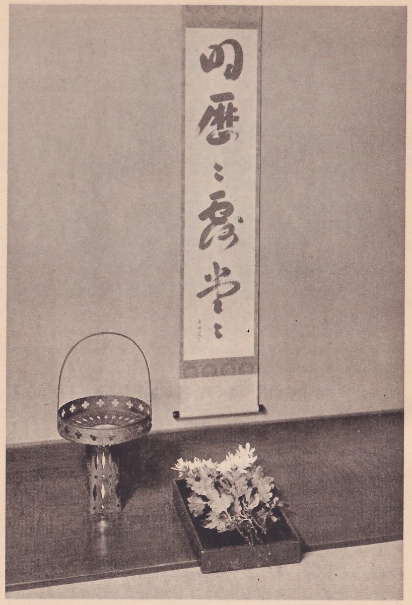 Photograph of an alcove with a hanging scroll of brush writing in large characters, 明歴々露堂々, a small vase shaped like a basket with a handle on a pierced stand beside it, and a dark tray of white flowers at the front.