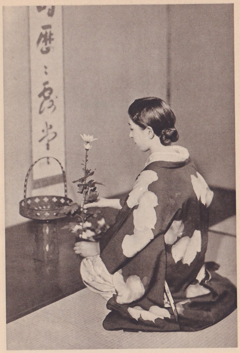 Photograph of a young woman in a dark kimono with large pale clouds, kneeling in profile facing left and holding a long stem with a white flower, with a small basket vase and a hanging scroll beside her.