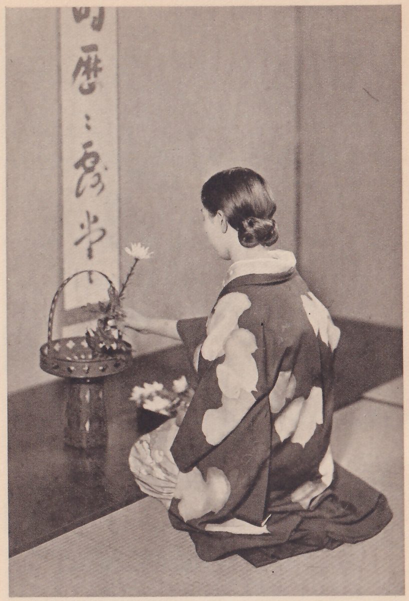 Photograph of the same young woman kneeling with her back to the camera at an alcove, reaching to place a white flower in a small basket vase with a handle; a dark flower tray is at her left and a hanging scroll of brush writing is on the wall.
