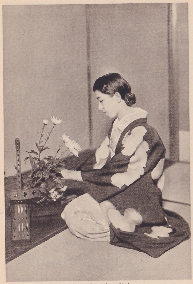 Photograph of the young woman kneeling in profile facing left and adding a branch to white flowers in a vase with a pierced stand, in a corner of a mat room.