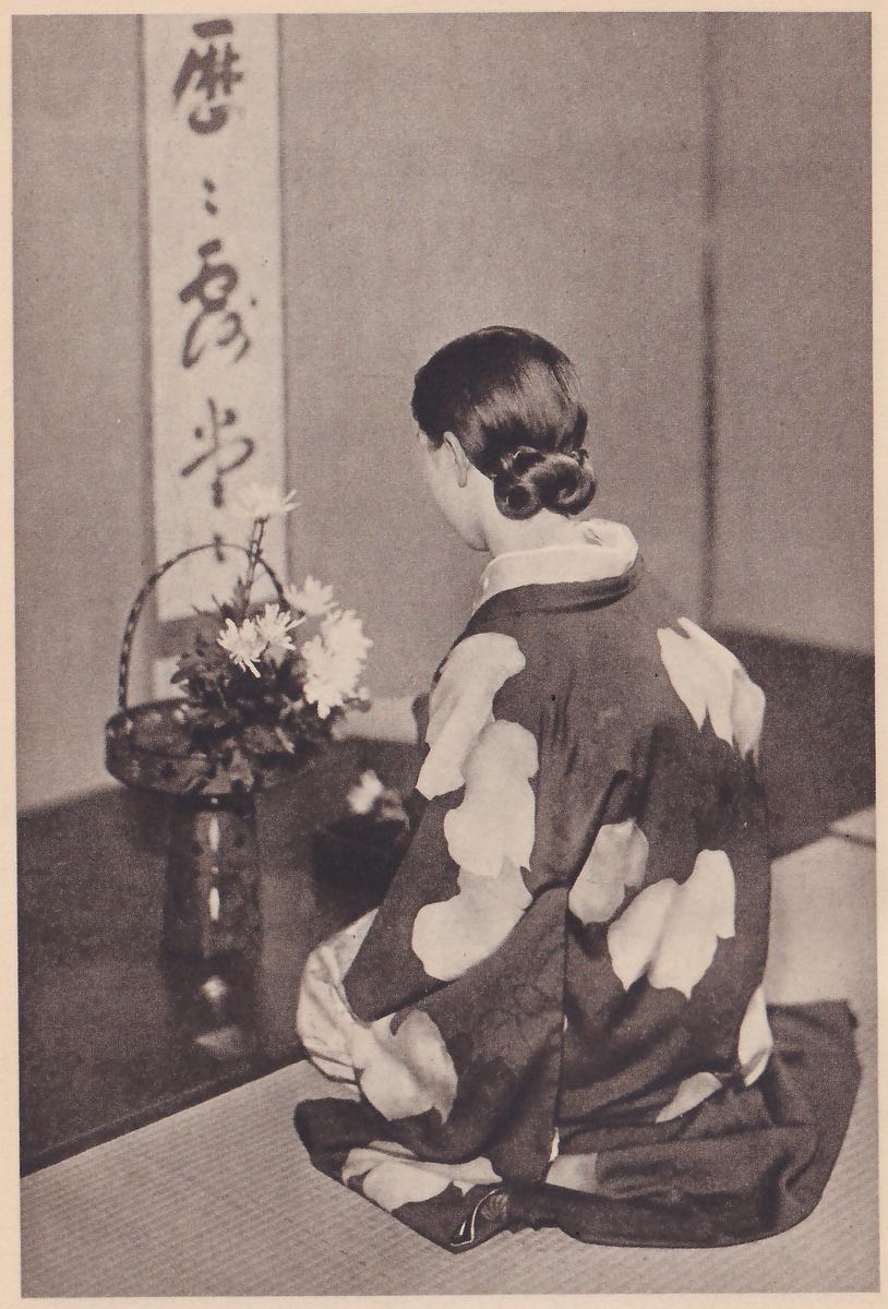 Photograph of the young woman kneeling with her back to the camera, in front of the basket vase, which now holds a fuller bunch of white flowers, with the hanging scroll above.