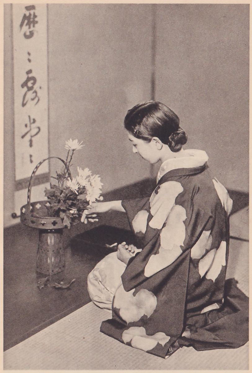 Photograph of the young woman kneeling in profile facing left, her right hand at a stem of the white flowers in the basket vase and her left hand in her lap; cut leaves lie on the floor.