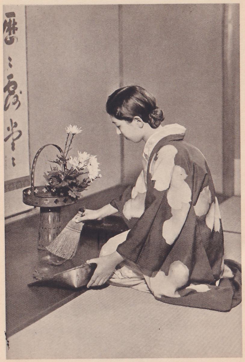 Photograph of the young woman kneeling in profile facing left, sweeping the floor in front of the finished arrangement of white flowers with a small broom into a dustpan she holds in her other hand.