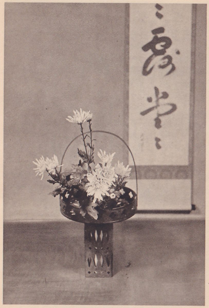 Photograph of the finished arrangement: white chrysanthemums of several sizes in a dark round vase with a handle, on a pierced tall stand, in front of a hanging scroll of brush writing.