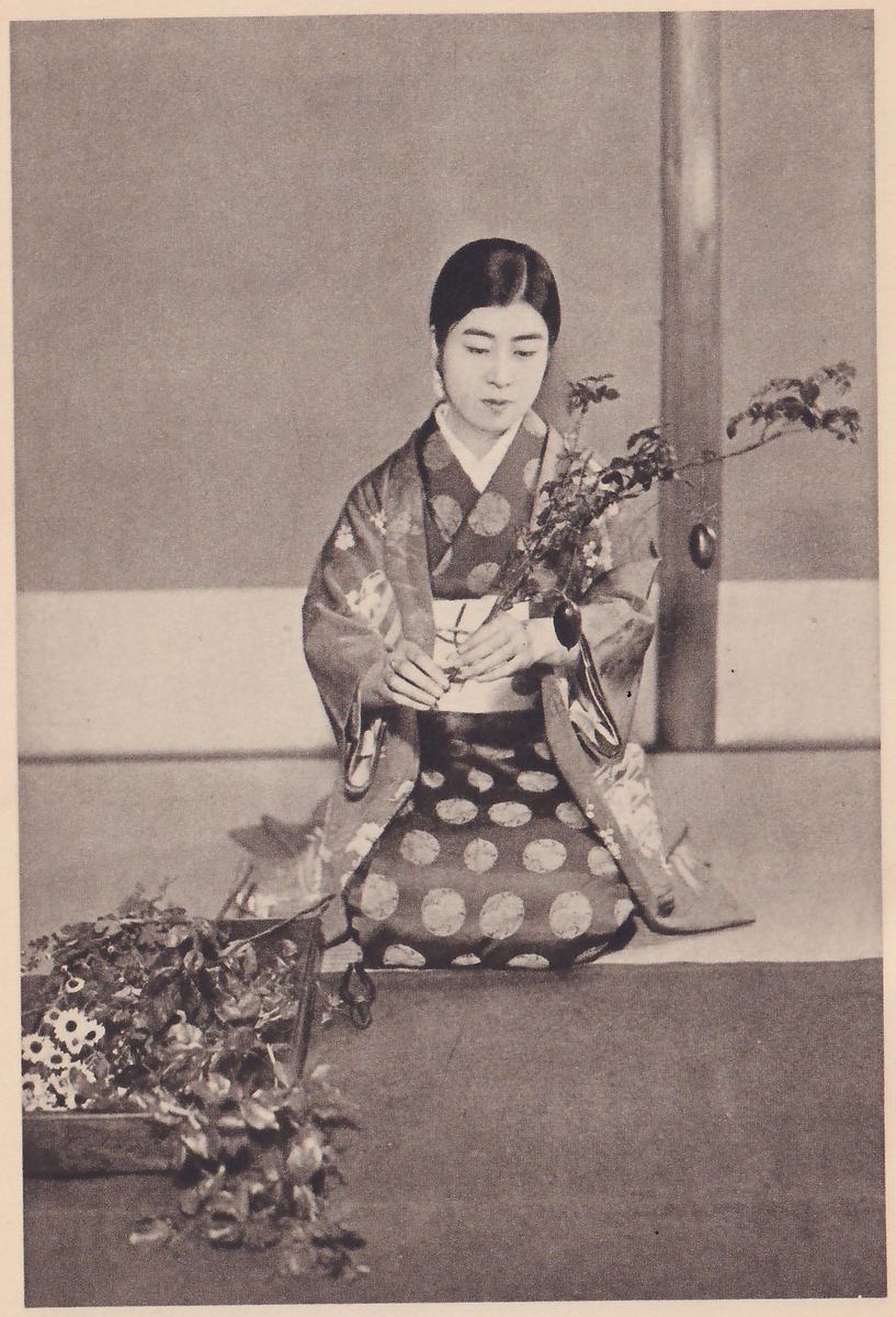 Photograph of a young woman in a kimono with a pattern of large dots on the skirt, kneeling in front of a wall and holding a leafy branch with one small hanging oval fruit in both hands; at the left a tray of flowers and leaves lies on the floor.