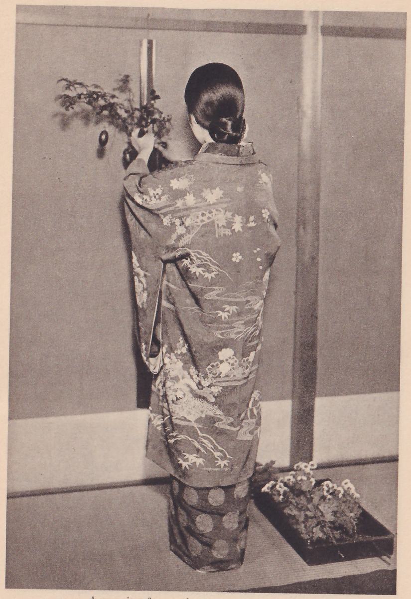 Photograph of a woman in a kimono with a pattern of maple leaves and water, seen from behind, standing in front of a wall and reaching up with one arm to a hanging bamboo vase on a post; a tray of flowers lies on the floor at the right.