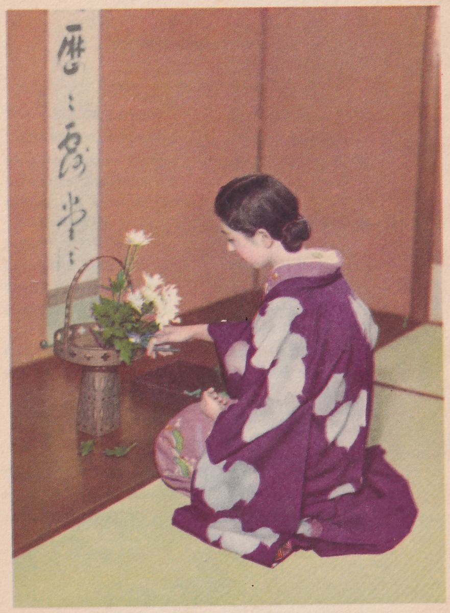 Color photograph of a young woman in a purple kimono with large pale clouds, kneeling on a green mat in the corner of a room and facing left, her right hand in the white flowers of a tall metal basket vase with a handle and her left hand in her lap; on the reddish-brown wall beyond the vase hangs a narrow scroll of brush writing.