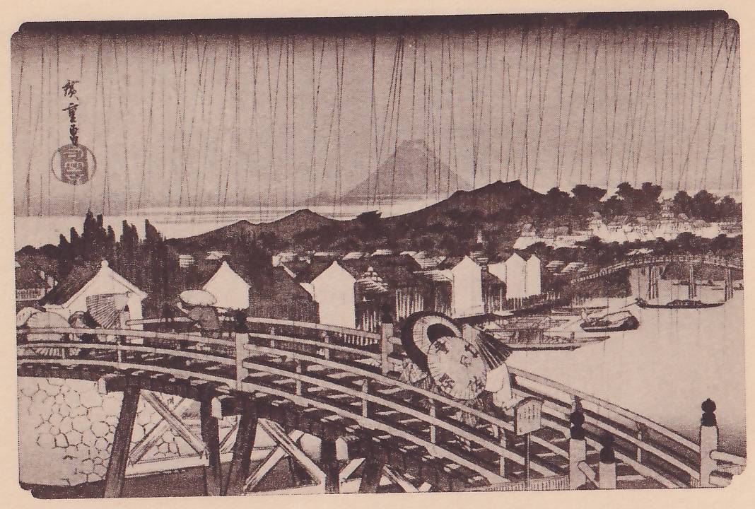 Print of a rainy day seen from a high arched wooden bridge: long thin lines of rain fall across the sky, a cone-shaped mountain rises above low hills in the distance, white-walled storehouses and roofs fill the middle ground, and a second long bridge and boats lie on the water at the right. People under umbrellas and wide hats cross the near bridge, and a small signboard stands on a post at the right. A signature and seal are at the upper left.
