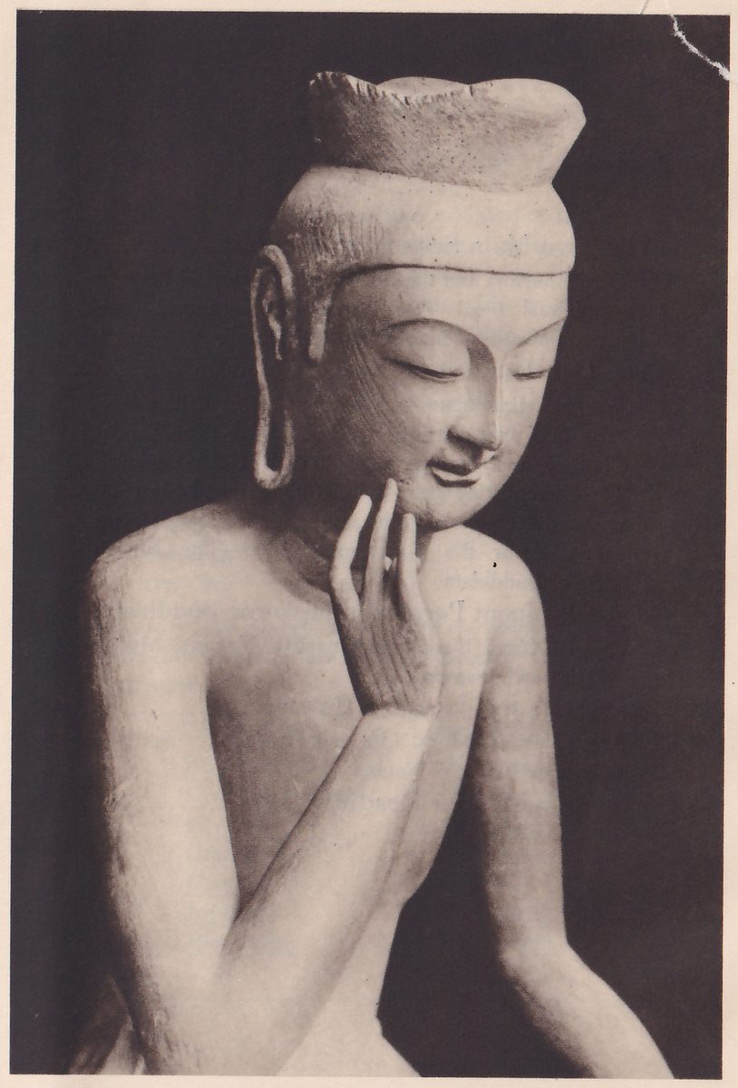 Photograph of a pale statue of a bare-chested figure, shown from the chest up on a dark ground, with a tall flat-topped headdress, long earlobes and lowered eyes; one hand is raised with its fingertips touching the cheek, and the other arm rests lower.