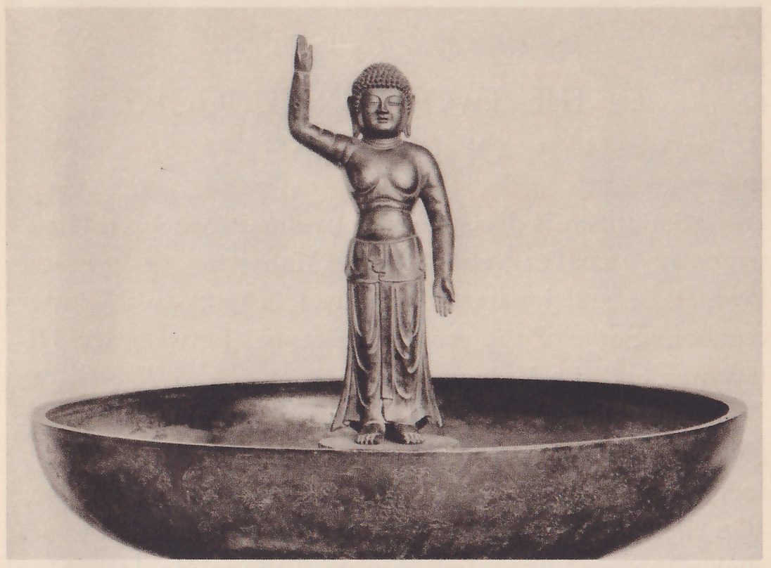 Photograph of a small statue of a standing figure in a long skirt, one arm raised straight up and the other held down at its side, standing barefoot in the middle of a very wide shallow round basin that fills the width of the picture.