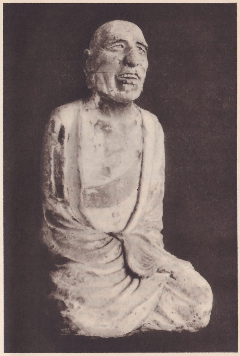 Photograph of a pale, chipped statue of a seated bald man with a deeply lined face and an open mouth, on a dark ground; a robe falls over his shoulders and lap, and his hands are hidden in its folds.