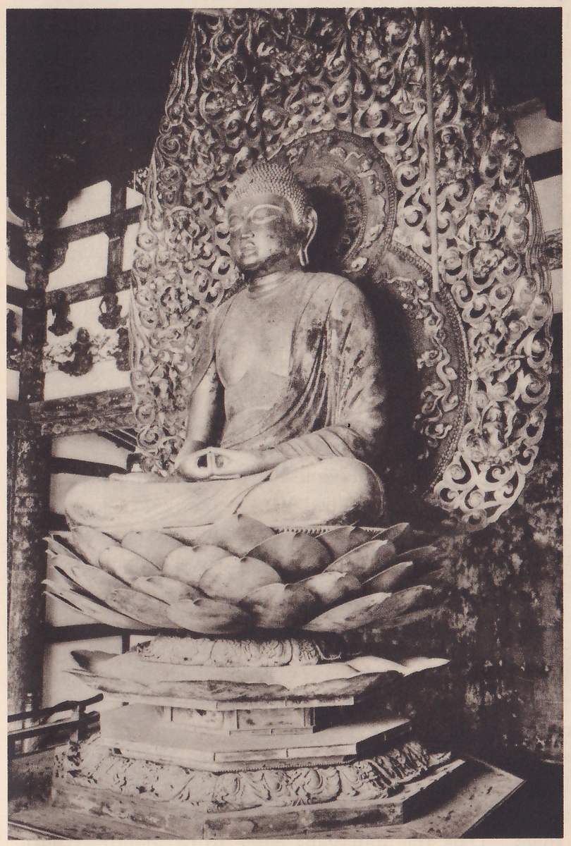 Photograph of a large statue seated cross-legged on a pedestal of lotus petals inside a wooden building, both hands joined in its lap; behind its head and shoulders rises a tall oval halo of swirling openwork.