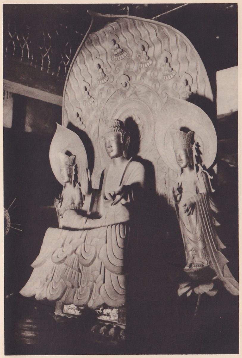 Photograph of three statues in a dark hall: a larger seated figure at the center with one hand held out low, a smaller figure at the left and a standing figure at the right, each with a halo; behind them rises a tall leaf-shaped back panel carved with wavy lines and small figures.