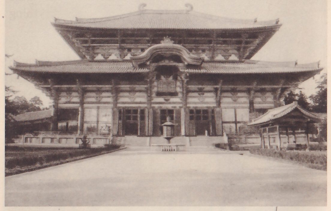Photograph across a wide open yard of the front of a very large wooden hall with stacked tiled roofs, the lower one with a curved gable over the center of the front; a row of tall posts holds up the lower roof, a lantern stands before the steps, and a small roofed building shows at the right edge.