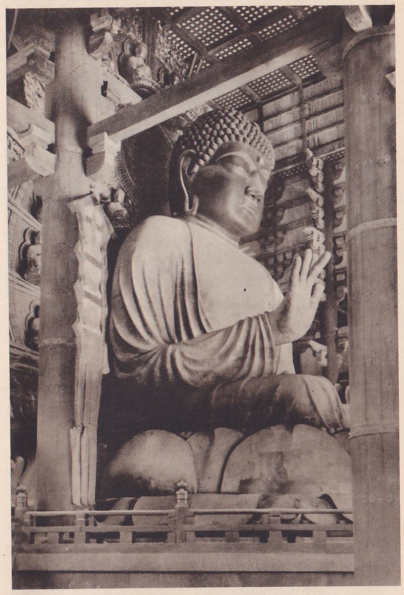 Photograph, taken from low down, of a very large seated statue with tightly curled hair in a dark wooden hall; the figure faces the viewer's right with one hand raised, and sits on a base of large rounded petals behind a low railing, with thick wooden posts at the left and right of the picture.