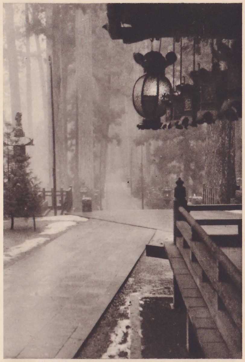 Photograph of a misty path among very tall straight trees: a wide paved walkway runs away from the viewer toward the center, a wooden railing lines the right side, and a large hanging lantern with a round body and smaller ones beside it hang from a dark eave at the upper right; patches of snow lie on the ground beside the walkway.