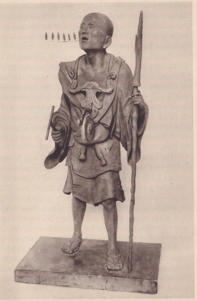 Photograph of a standing statue of a lean bald man in a short robe and straw sandals, his face tilted up and his mouth open; he holds a tall staff with a spear-like head in one hand at the right, a round object hangs on his chest from straps, and a row of tiny upright figures extends from his open mouth toward the viewer's left; he stands on a low rectangular base.