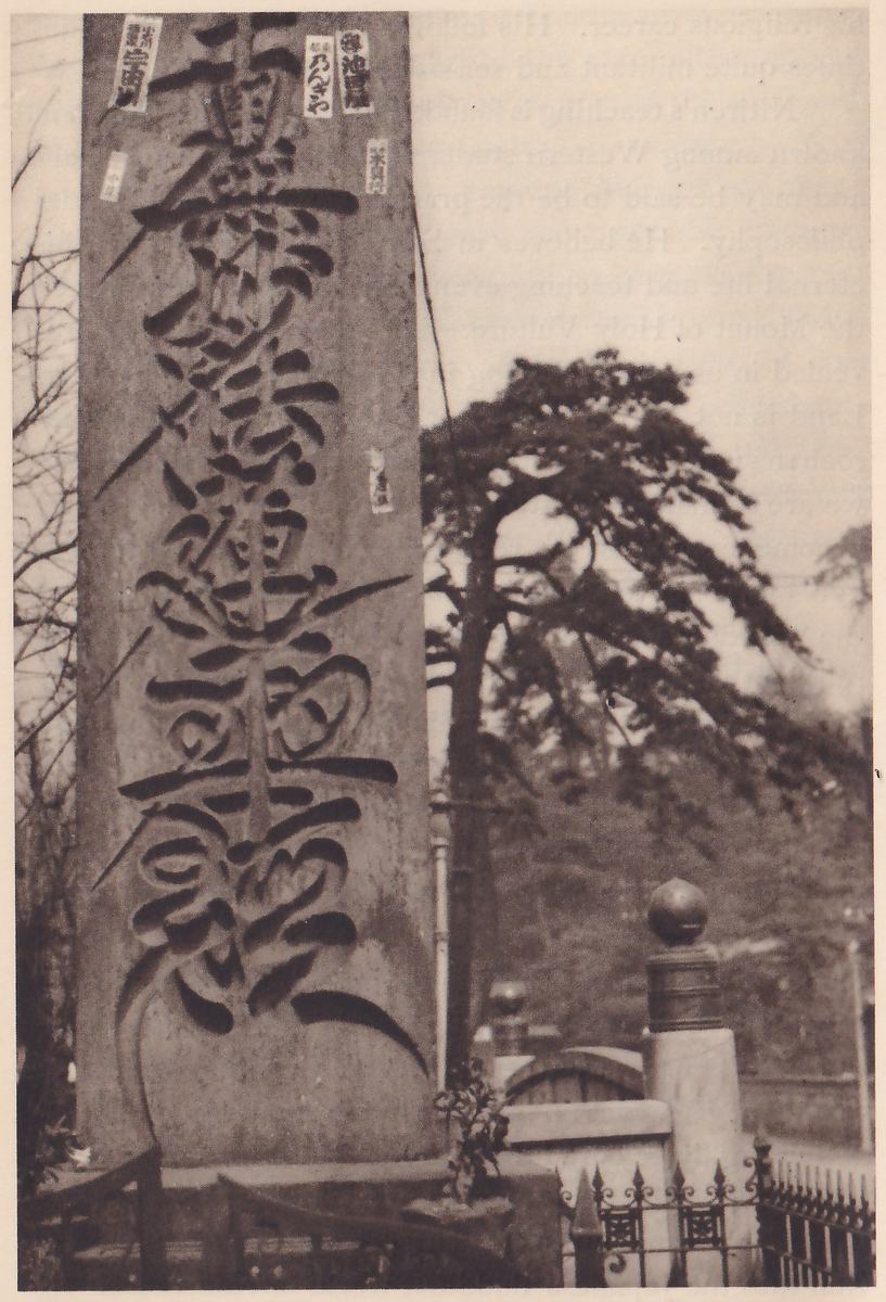 Photograph of a tall rough stone slab with a large inscription carved down its face in one column of heavy brush-shaped characters, small paper slips stuck on its upper part; behind it at the right stand a pine tree, a low wall and an iron fence with a stone post.