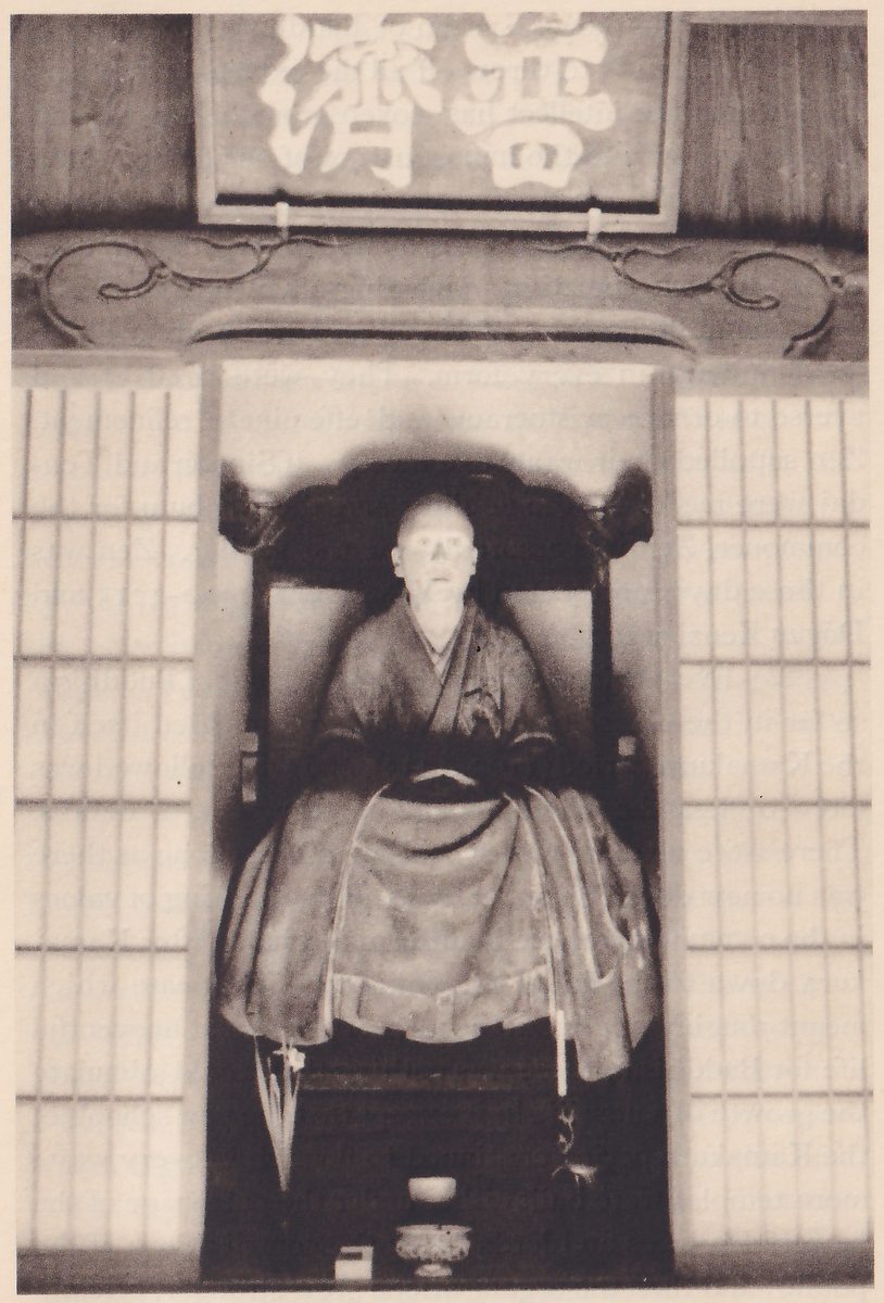Photograph of a seated statue of a bald figure in layered robes, hands resting in the lap, on a high-backed dark chair inside a narrow recess between two paper sliding screens; above the recess a wooden board shows two large pale characters, Fusai, one of Musō's titles as national teacher, and a small dish stands on the floor in front.