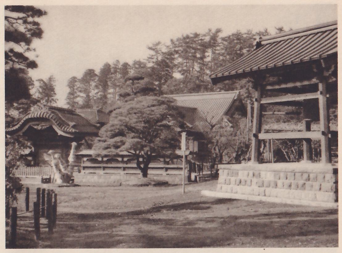 Photograph of a temple yard: at the right an open wooden framework under a tiled roof stands on a raised stone base; across the open ground behind it are a pine tree with a rounded top, a low building with a curved roof at the left, and a line of pines along the horizon, with a row of short posts at the left edge.
