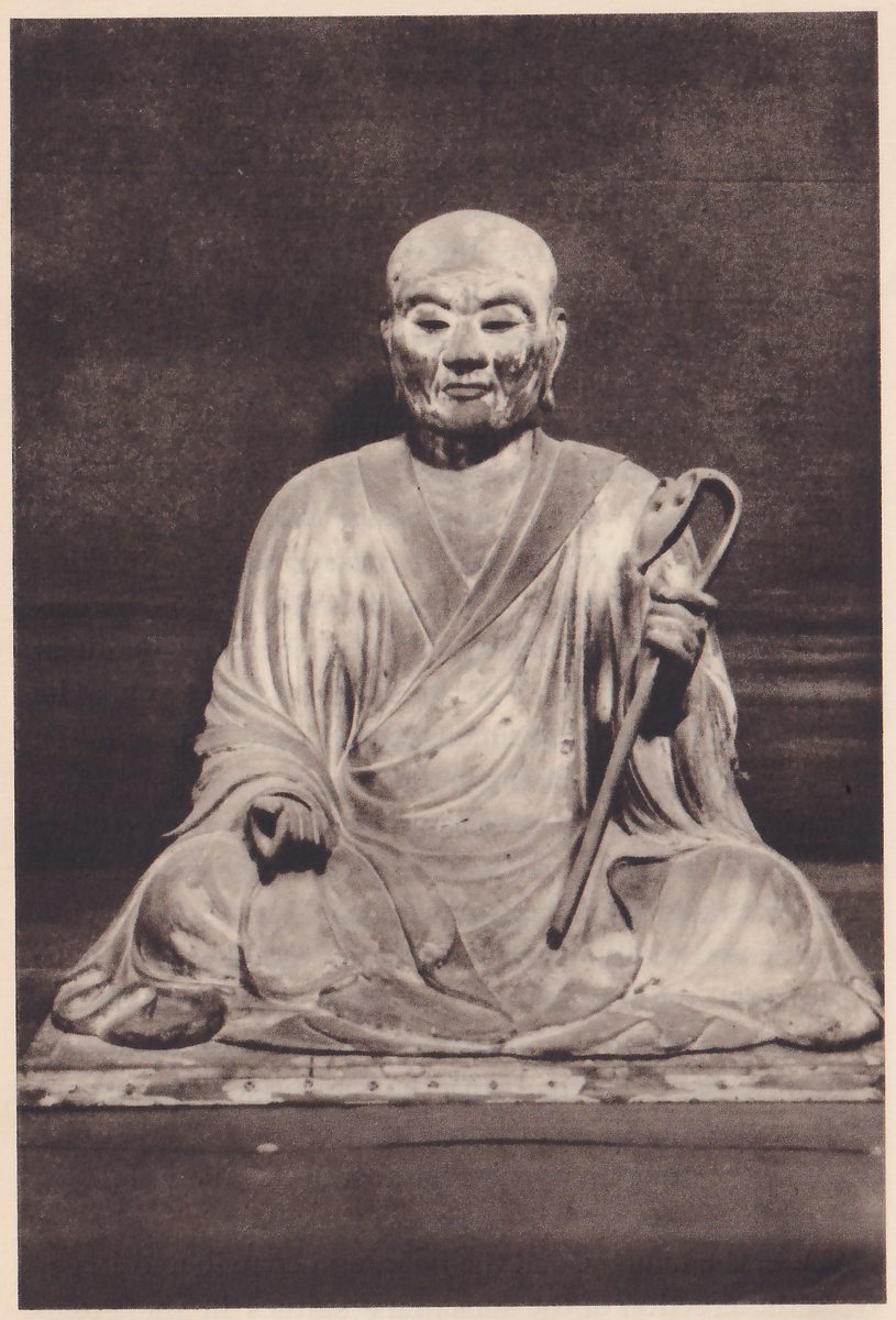 Photograph of a seated statue of an elderly bald man with a lined face, in a robe that crosses over the chest, seated cross-legged on a low platform on a dark ground; one hand at the viewer's right holds a staff with a looped top, and the other hand rests on the knee at the left.