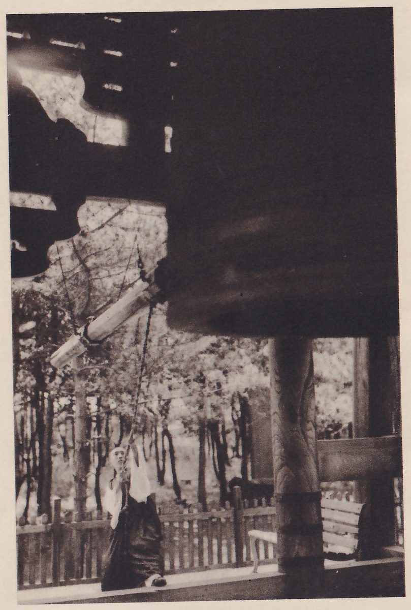 Photograph taken from under the roof of a bell tower: a huge dark bell fills the upper right, a long pale beam hangs at a slant on ropes in front of it, and a person in a dark skirt and pale top stands at the lower left beyond the beam, with trees and a low wooden fence beyond the open sides.