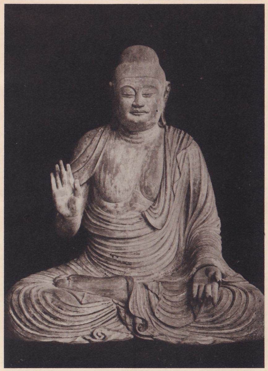 Photograph of a seated statue with lowered eyes, a rounded topknot and a robe open across the chest, sitting cross-legged on a dark ground; one hand is raised at the viewer's left with the palm toward the viewer, and the other rests on the knee at the right.