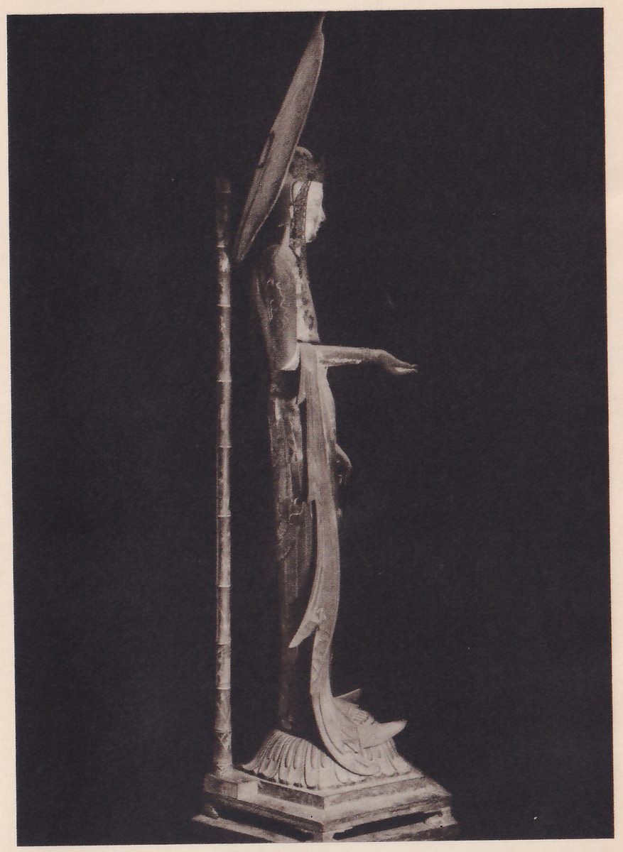 Photograph of a tall slender statue seen from the side in a dark room, standing on a stepped base and facing right with one arm stretched forward; behind its head rises a tall leaf-shaped halo, and a thin tall jointed rod stands at the left.