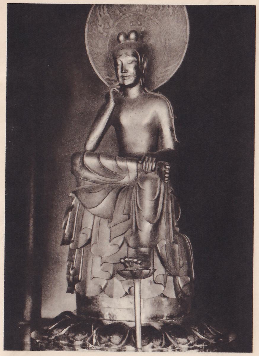 Photograph of a statue seated on a raised seat with one leg crossed over the other knee, the head leaning on a hand raised to the cheek, a round halo behind the head and a pedestal of lotus petals below; the other hand rests on the crossed leg, and the lowered foot rests on a small lotus on a thin stem.