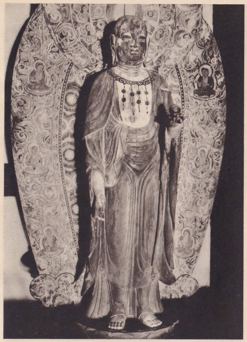 Photograph of a standing statue in a long robe with a necklace-like ornament on the chest, one hand raised at the viewer's right holding a small round object and the other hanging at the side, standing barefoot on a round base in front of a tall pointed-oval backing panel painted with swirling patterns and small seated figures.