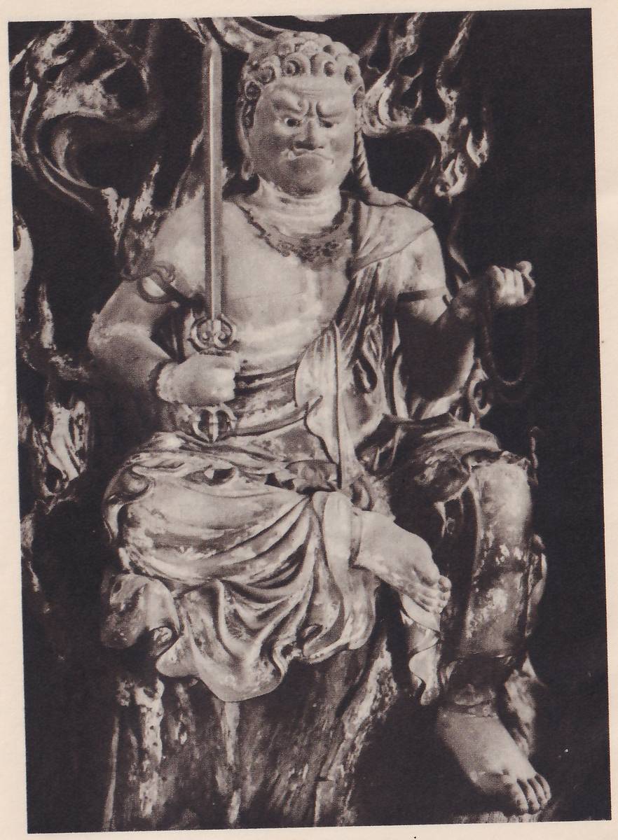Photograph of a seated statue of a fierce-faced figure with curly hair, a frowning brow and bared teeth, holding a long upright sword in the hand at the viewer's left and a rope in the raised hand at the right, one leg drawn up and the other foot hanging down; flames and rough rock shapes rise behind it.