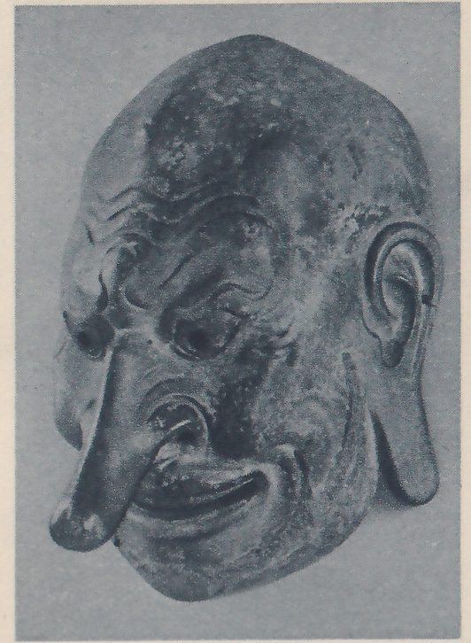 Photograph of a carved mask turned a little toward the viewer's left: a rounded head with no hair showing and wrinkles across the forehead, small dark eyes, a very long nose that droops toward the lower left, a wide mouth turned up at the corners, and a large ear with a long lobe at the right. The surface is dark and worn.