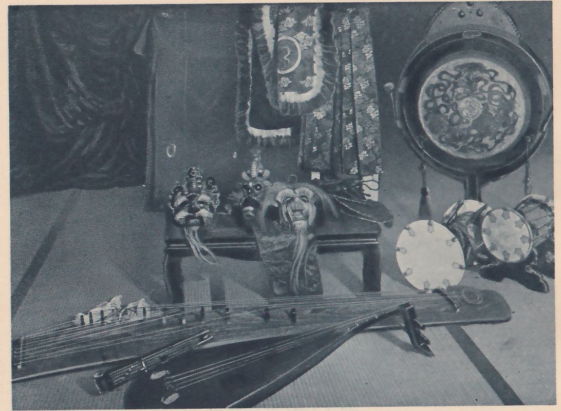 Photograph of objects arranged on a mat floor in front of a dark drape: a low table at the center holds carved masks with long strands hanging from them, behind which hang long patterned cloths and a bordered robe. At the right a large round drum in a frame stands with a tassel hanging below it and smaller round drums beside it, and across the front lie long wooden stringed instruments with raised bridges, one overlapping the other.