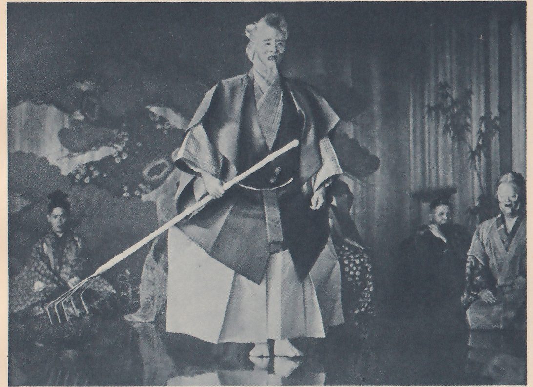 Photograph of a person standing at the center on a glossy floor in a loose dark coat with wide sleeves over a pale robe and wide pale trousers, a long rake slanted across the body in one hand; the face is pale, with an open mouth. At the left a person in a patterned robe kneels, and at the right more people kneel in shadow, one with a pale face. Behind them a painted backdrop of branches with small flowers shows at the left and a bamboo plant stands before striped panels at the right.