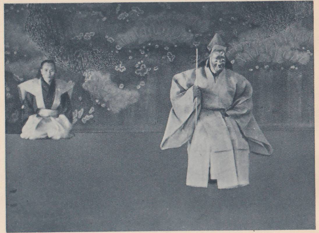 Photograph of two people on a bare floor in front of a dark painted backdrop with small white flowers. At the right a person stands in a wide-sleeved pale robe and a tall pointed cap, the face pale and showing little detail, holding a thin pole upright. At the left a person kneels in a pale jacket with wide shoulders and white wide trousers, facing the viewer.