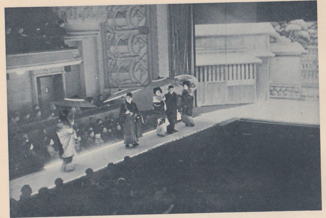 Photograph taken from above of a long pale raised walkway running diagonally from the lower left toward the upper right past rows of seated people, with a line of people in long robes walking along it, some holding open umbrellas. At the right end a pale two-storey building front with white-topped roofs and a low wall with a stone pattern stands in front of a dark curtain, and at the upper left a carved ornamental panel and a doorway show. Rows of heads fill the lower foreground.