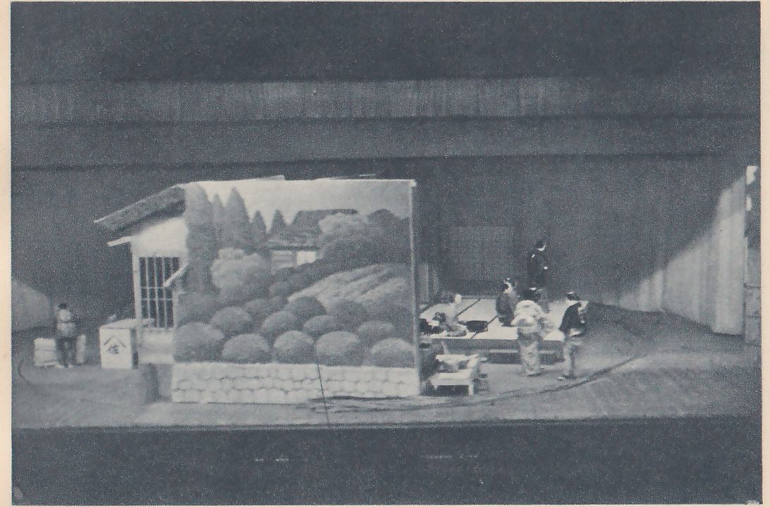 Photograph from a distance of a wide dark space with a lit set across the middle. At the left a small house with a tiled eave and a barred window stands beside a painted panel of a hillside with rows of round bushes and a roof, a low wall with a stone pattern along its foot. At the right several small figures stand and sit on a raised mat floor beside a low table, one more figure stands at the far left, and a tall pale wall rises at the right edge.