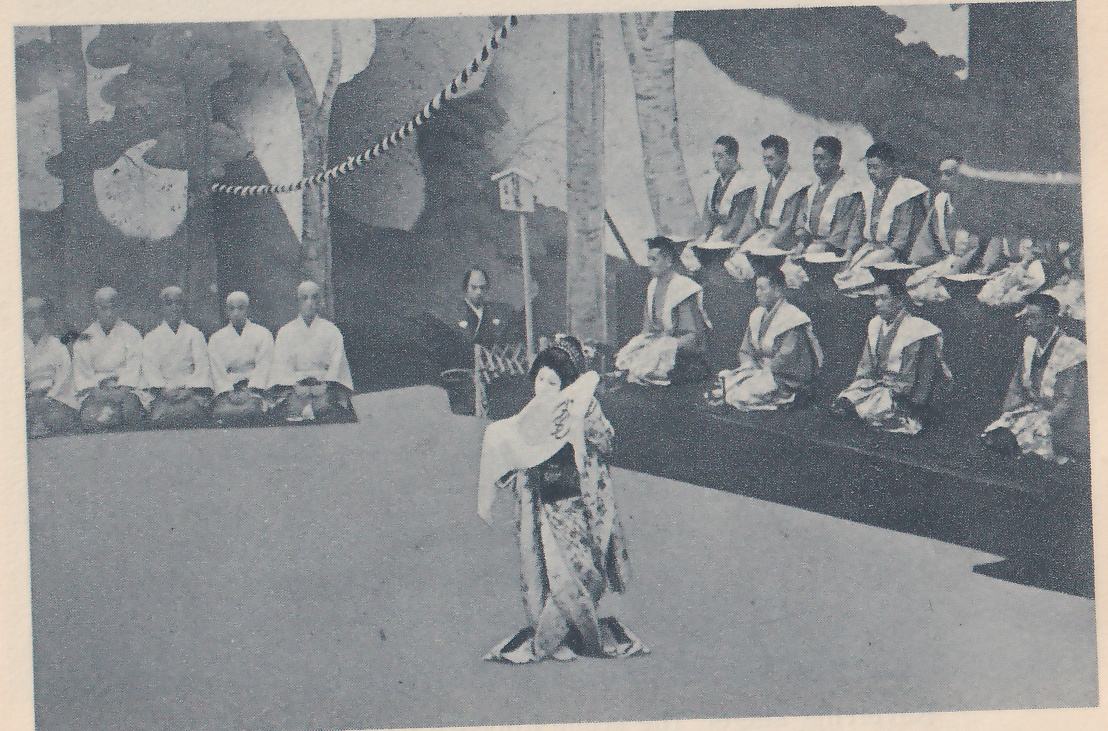 Photograph of a person in a long patterned robe with a wide sash standing alone at the center of a pale floor, holding a white fan raised in front of the chest. Behind them a row of men in white jackets and dark trousers sits at the left, and rows of men in pale wide-shouldered jackets sit on a dark raised tier at the right; a man in dark clothes sits behind a low fence at the center. A painted backdrop of tree trunks and branches fills the back, with a thick twisted rope hanging across it at the upper left and a small signboard on a pole near the center.