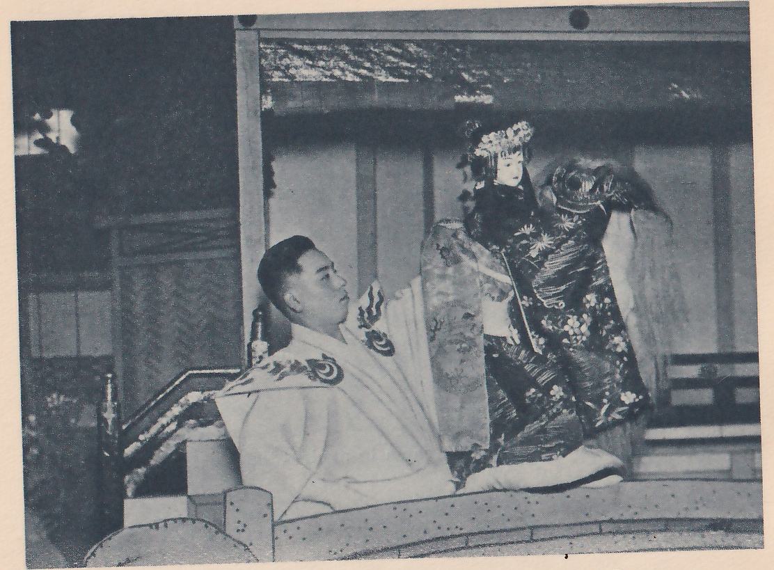 Photograph of a man in a pale robe with dark curling patterns on the shoulders, seated at the left in profile and looking toward the right, one arm raised at the side of a doll he holds up at the right. The doll has a pale painted face and a flowered headdress and wears a dark robe patterned with white flowers, a long sleeve hanging at its left. Behind them are paneled sliding doors, and a rail with small dots runs across the foreground.