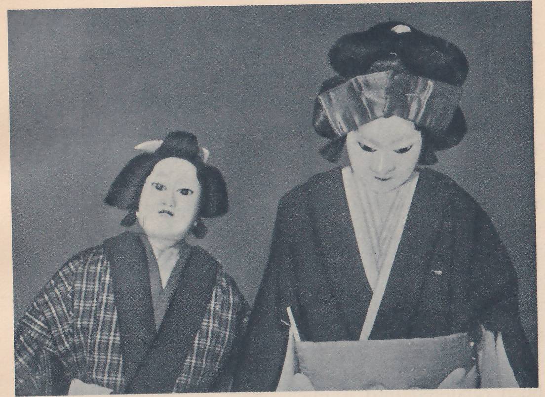Photograph of two figures with pale painted faces against a plain gray ground. The smaller at the left has dark hair swept into a bun tied with white bands and wears a plaid robe with a dark collar. The larger at the right has the eyes cast down, dark hair piled high with a shiny wide band around it, and a dark jacket over a white collar, and holds a pale rectangular object in front of the body.