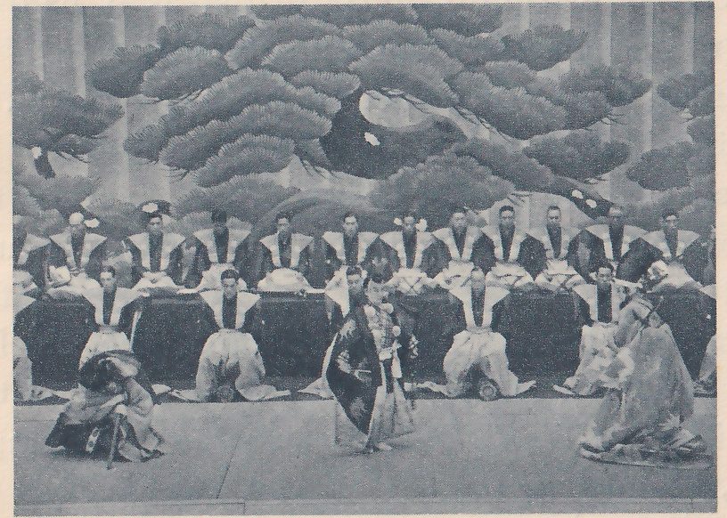 Photograph of a wide bare floor in front of a large painted pine with layered branches. Rows of men in pale wide-shouldered jackets and dark robes kneel and sit behind long dark low stands across the back. In front, three costumed figures stand and bend on the floor: at the left one crouches low with a sword at the side, at the center one stands in a long dark patterned coat, and at the right one bends forward in a pale patterned robe.