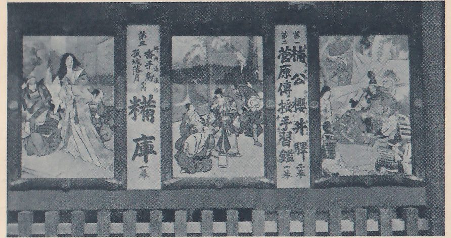 Photograph of the front of a dark wooden building with three large framed painted pictures hung side by side, separated by two tall narrow white boards of large vertical writing that list the bill: on the right board, first 楠公櫻井驛 in two acts and second 菅原傳授手習鑑 in one act; on the left board, third 孤城落月, its 糒庫 act. A fence with square posts runs along the bottom. The picture at the left shows a woman with long hair in a long pale robe with two armored men crouching beside her; the one at the center shows several men in robes gathered around a bottle on the floor; the one at the right shows a seated figure in armor beside a standing figure in a long robe at the left edge.