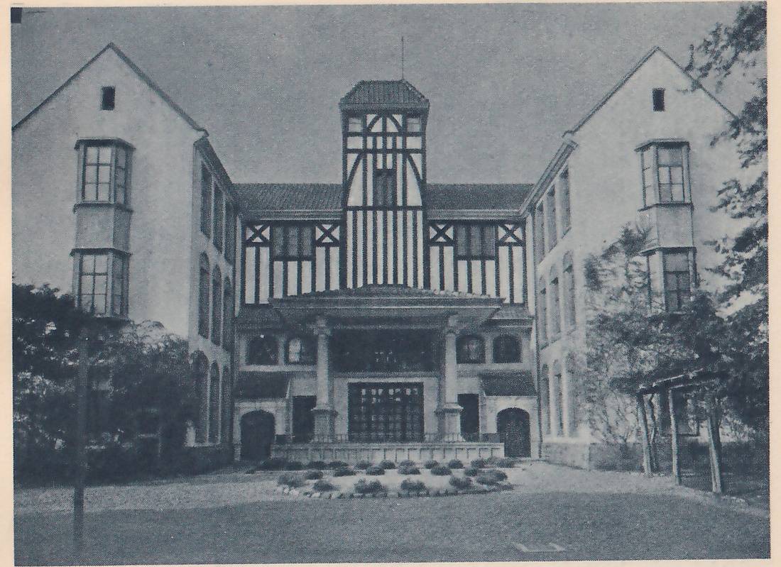 Photograph of the front of a large building seen straight on across a lawn: two tall pale gabled wings with bay windows stand at the left and right, joined by a lower center part with a tiled roof, a tower-like gable of dark timber framing and pale panels, and a porch with two round columns in front of a large many-paned window. Trees stand at the left and right, a small wooden frame like an arbor stands at the right, and a bed of low shrubs sits on the gravel in front.
