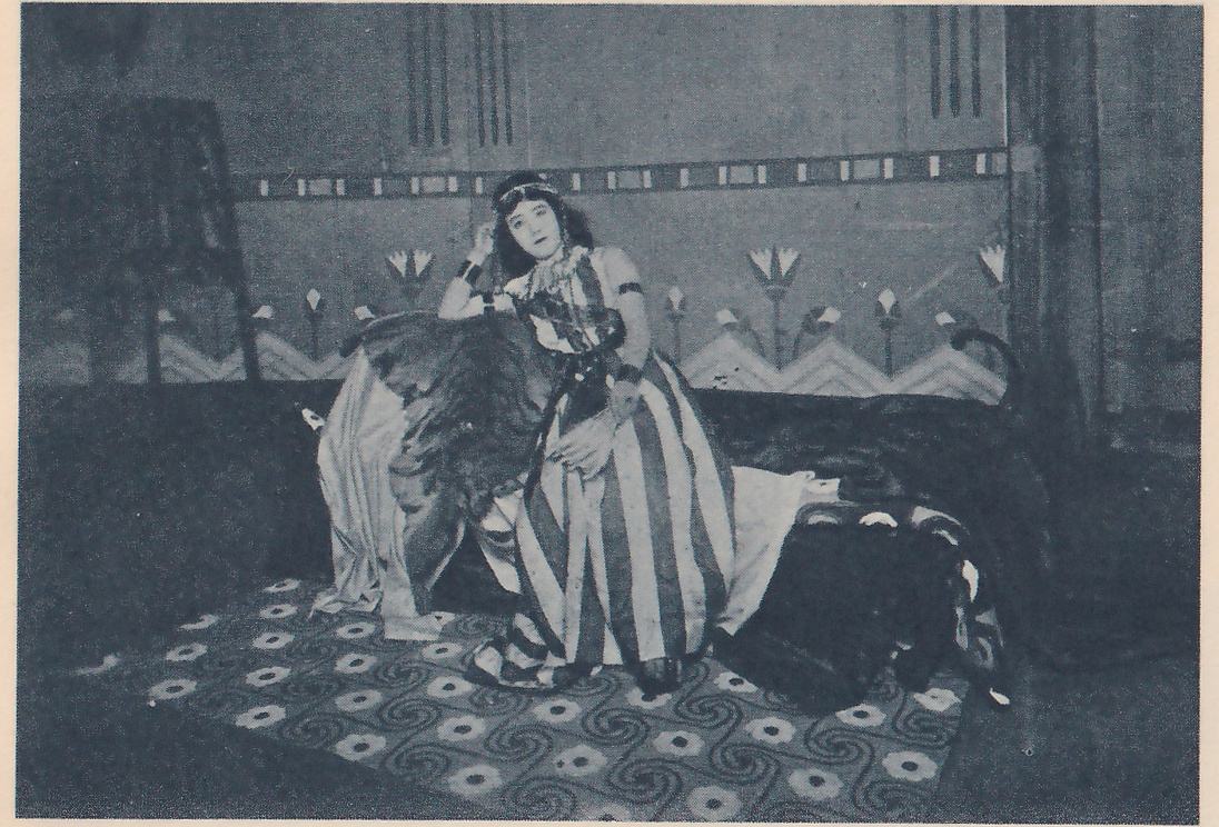 Photograph of a woman with dark wavy hair, a headband and beaded necklaces sitting on a low couch heaped with pale cloth and shaggy dark fur, leaning on one raised arm with the hand at her head and looking toward the viewer. Her long gown has wide vertical stripes in light and dark, and dark bands circle her upper arms. Behind her is a painted wall with a row of tall flower shapes above a zigzag band and a strip of small rectangles at the top, and the floor carpet is patterned with spirals and round flowers.