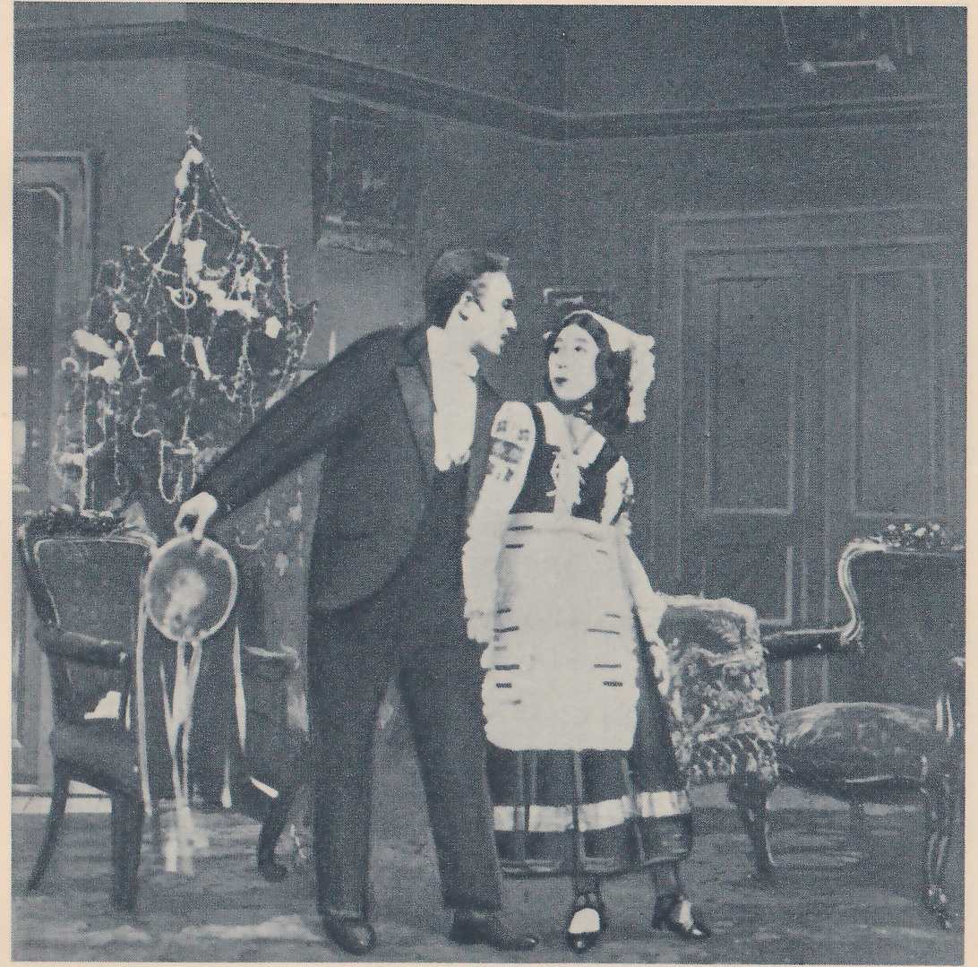 Photograph of two people in a furnished room. A man in a dark suit and a high white collar leans toward a young woman, his mouth open, with one arm stretched down at his side holding a round flat object with ribbons hanging from it; she stands upright in a white cap, a dark bodice with embroidered sleeves and a white apron with dark stripes, looking up at him. At the left a conical tree hung with strings and small ornaments stands behind a carved chair, more chairs stand at the right, and paneled doors and a framed picture are on the wall behind.